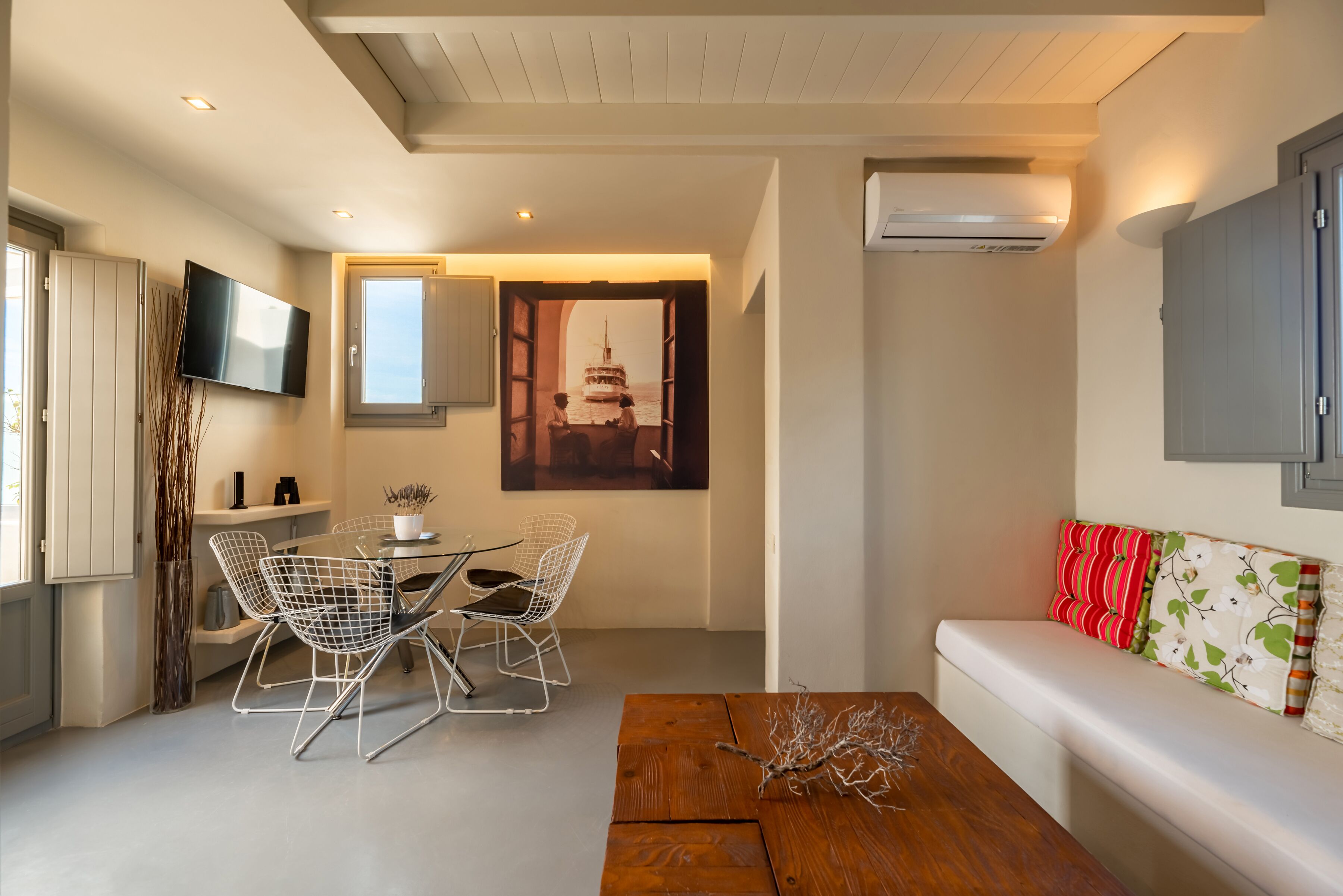 A modern living room with a wooden coffee table, a white sofa with patterned cushions, a dining area with a glass table and wire chairs, a mounted TV, and an air conditioning unit.