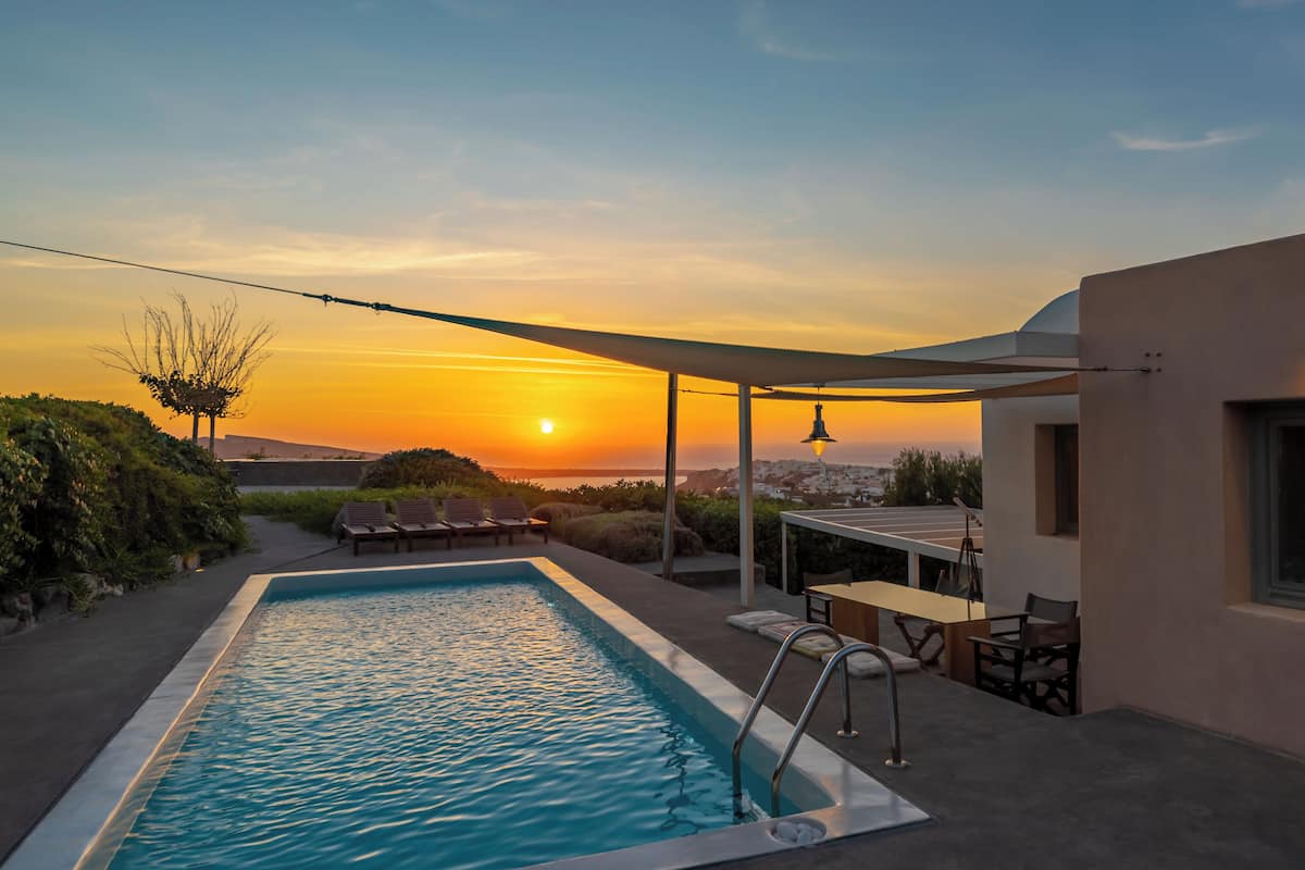 A swimming pool with lounge chairs and a table, overlooking a sunset with a view of a distant town.