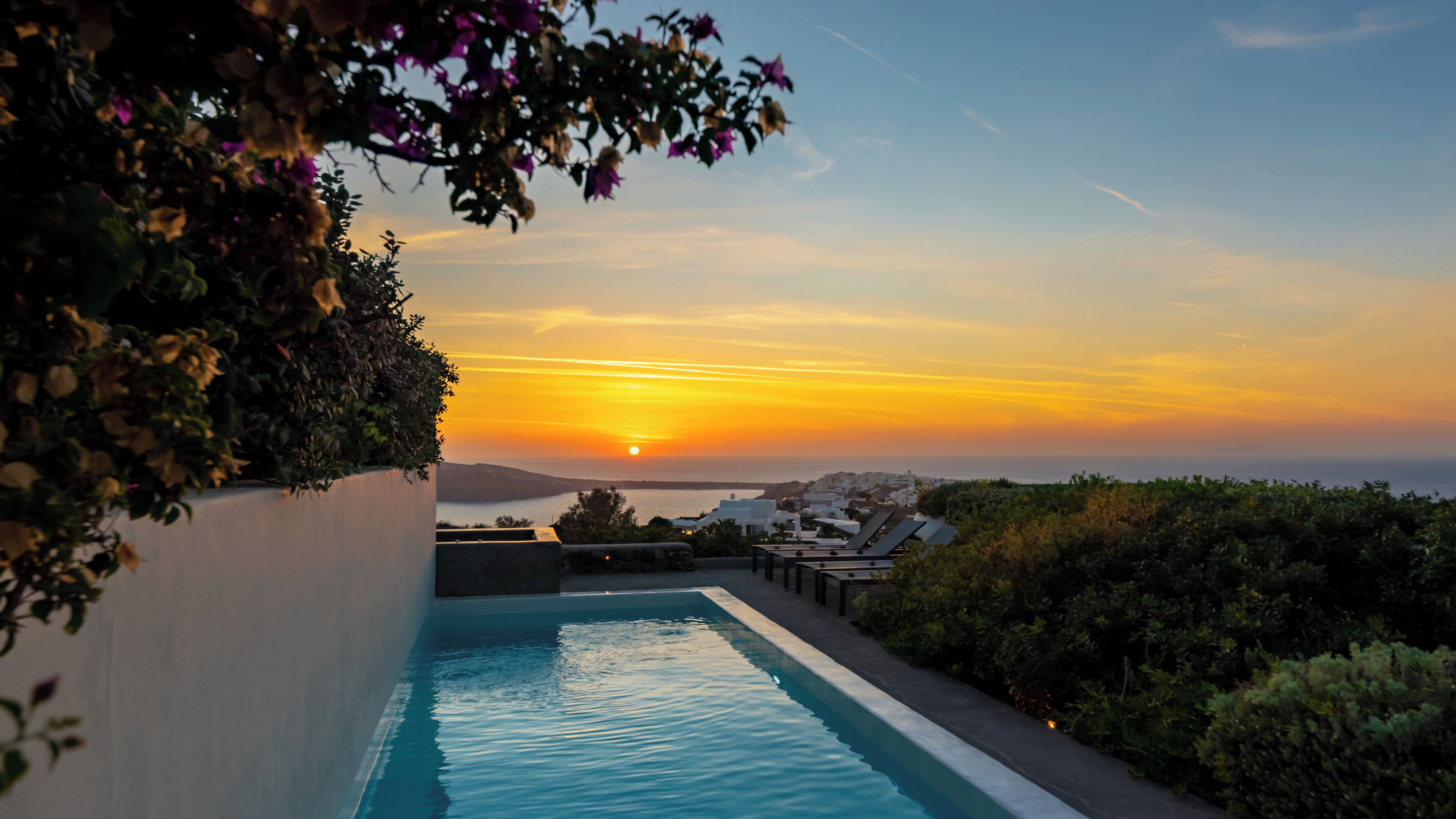A swimming pool with a sunset view, surrounded by greenery and overlooking a coastal area.