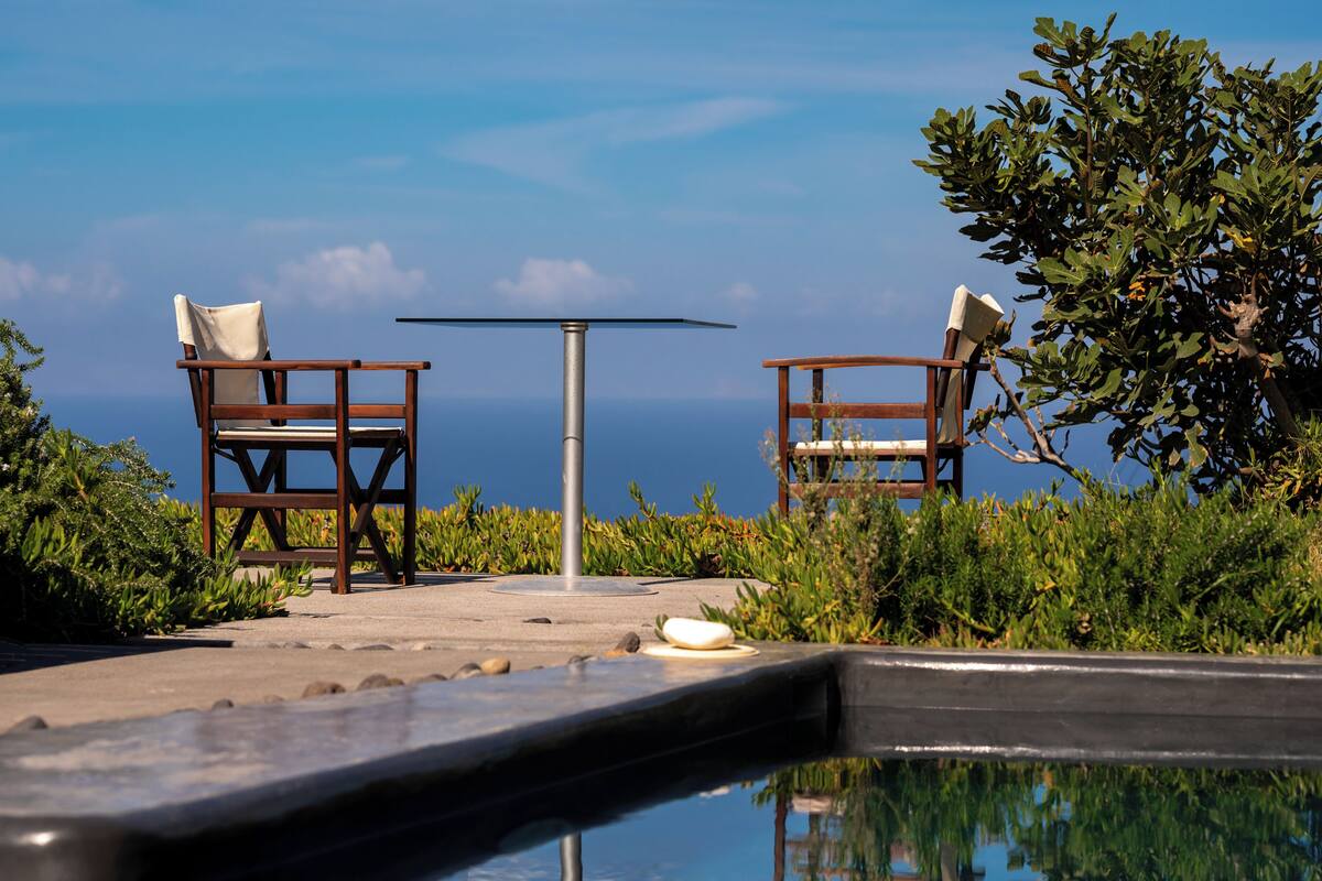 A poolside area with two wooden chairs and a table, surrounded by greenery and overlooking a body of water.