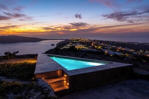 A rectangular pool with steps on one side, surrounded by a rocky landscape and overlooking a coastal town at sunset. - 270 Oia's View (Santorini)