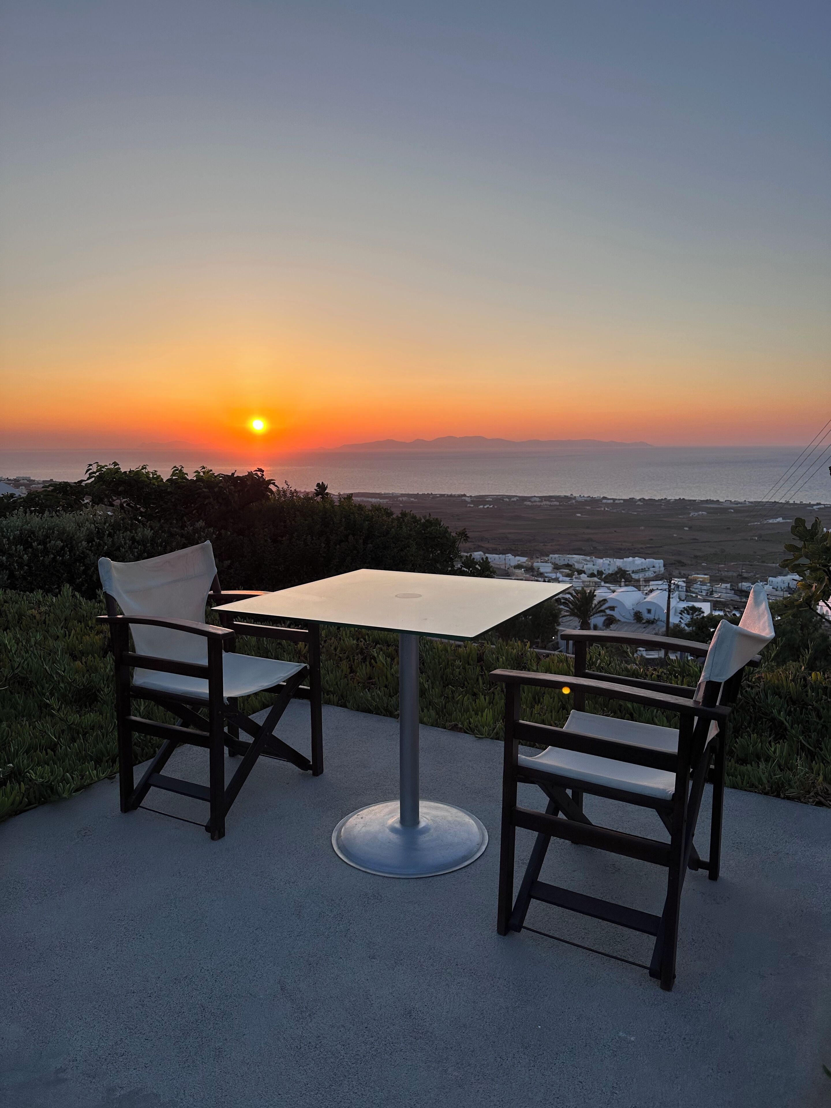 Ein Sitzbereich im Freien mit Tisch und Stühlen mit Blick auf einen Sonnenuntergang über dem Meer.