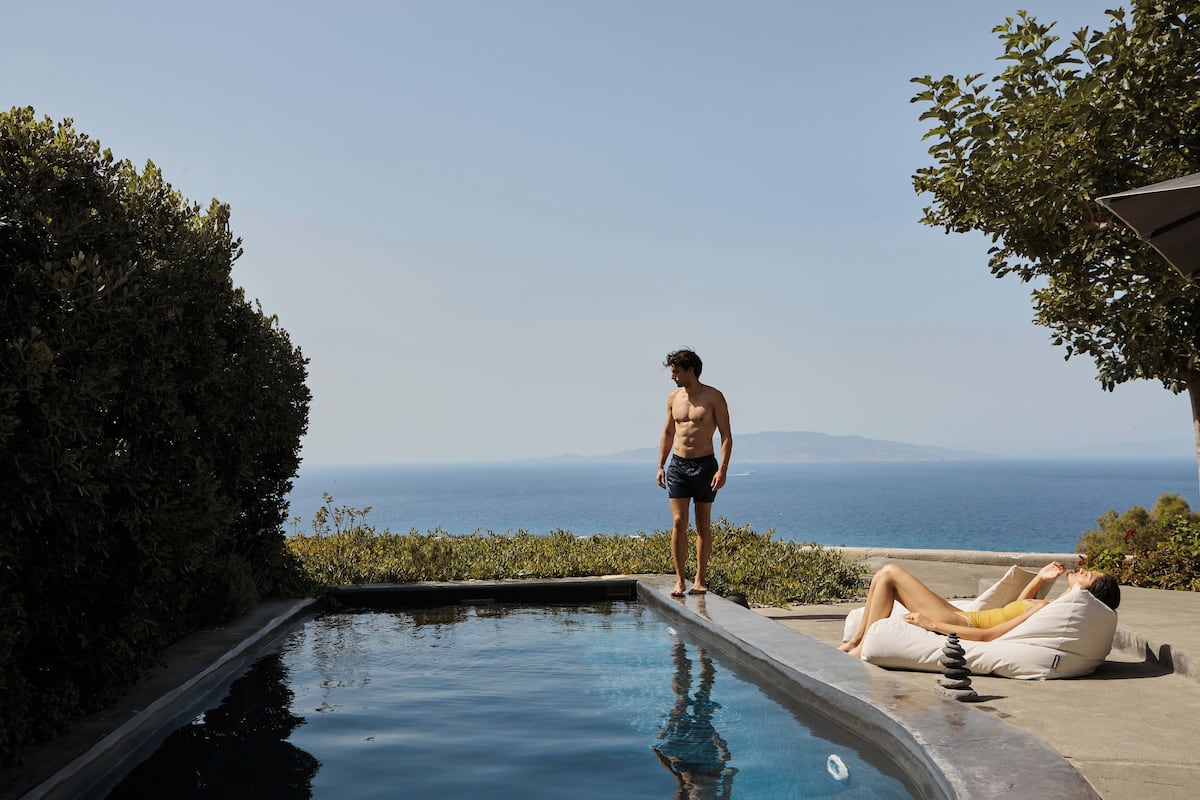 A man standing by a pool with a view of the sea and a woman relaxing on a lounge chair.