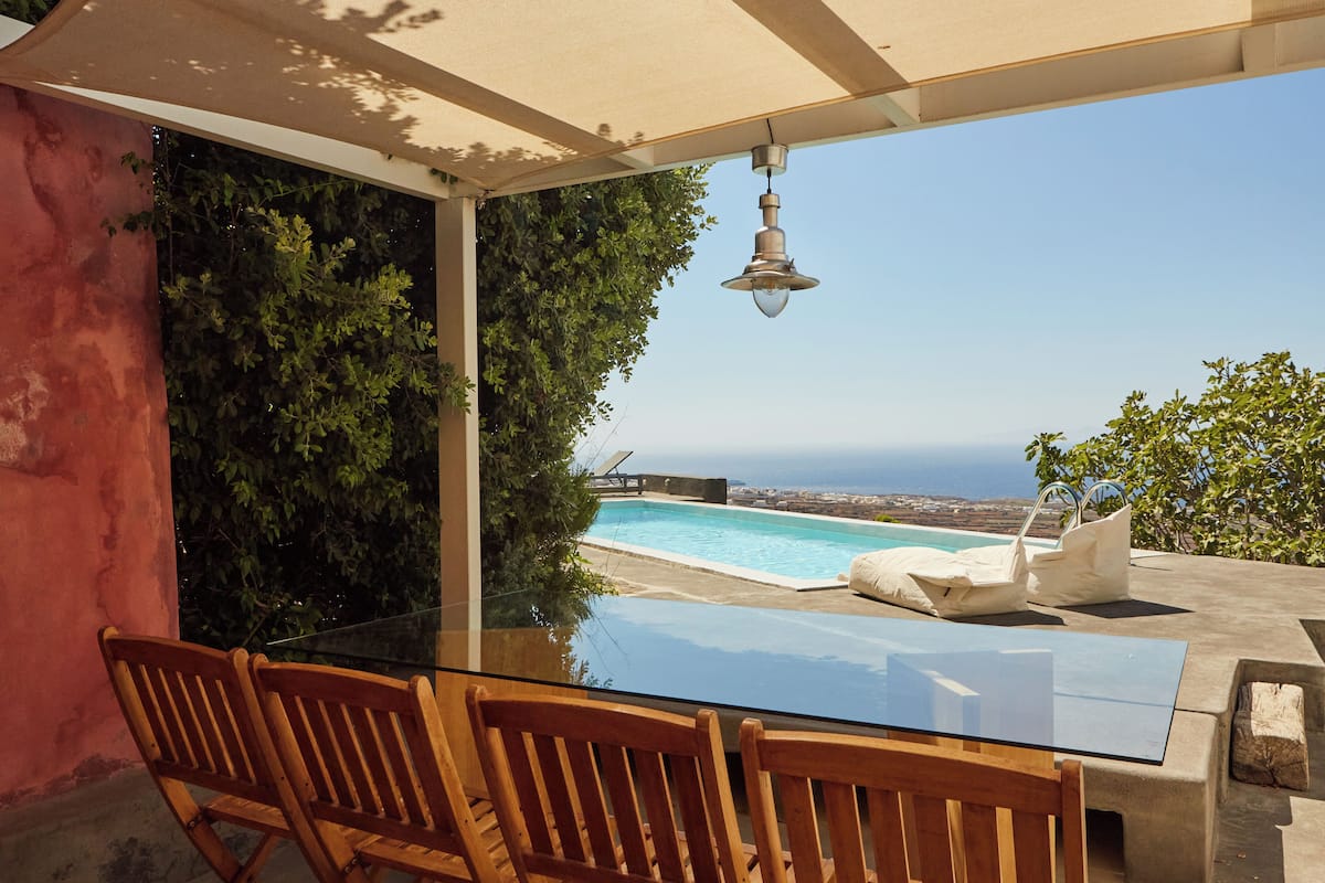 A glass table with wooden chairs, a hanging light fixture, and a view of a swimming pool and ocean.