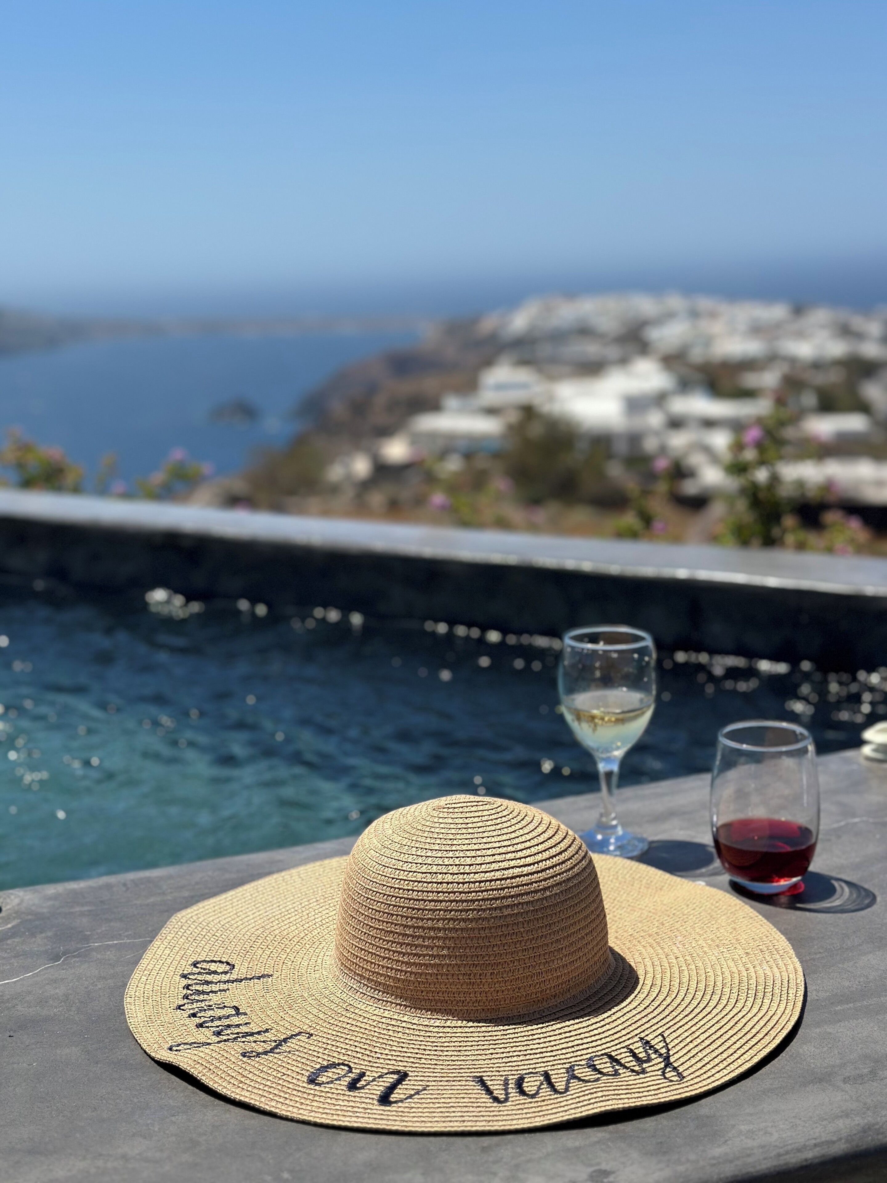 A straw hat with "Enjoy your vacation" written on it, two wine glasses, one with white wine and the other with red wine, and a view of a coastal town with buildings and a body of water.