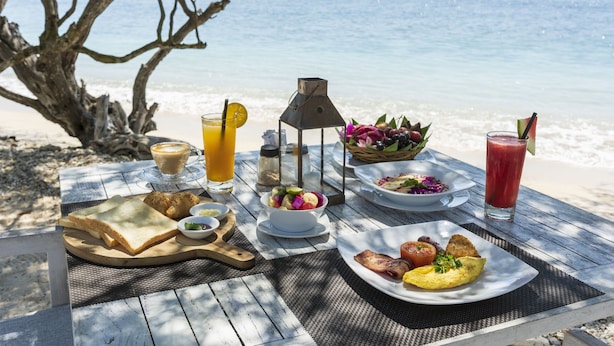 Ett frukostbord vid stranden med ett varierat utbud av mat och dryck, inklusive rostat bröd, ägg och en sallad, med havet som bakgrund.