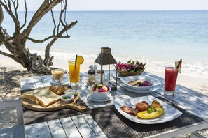 Una mesa de desayuno junto a la playa con una variedad de alimentos y bebidas, incluyendo tostadas, huevos y una ensalada, con el océano como telón de fondo.