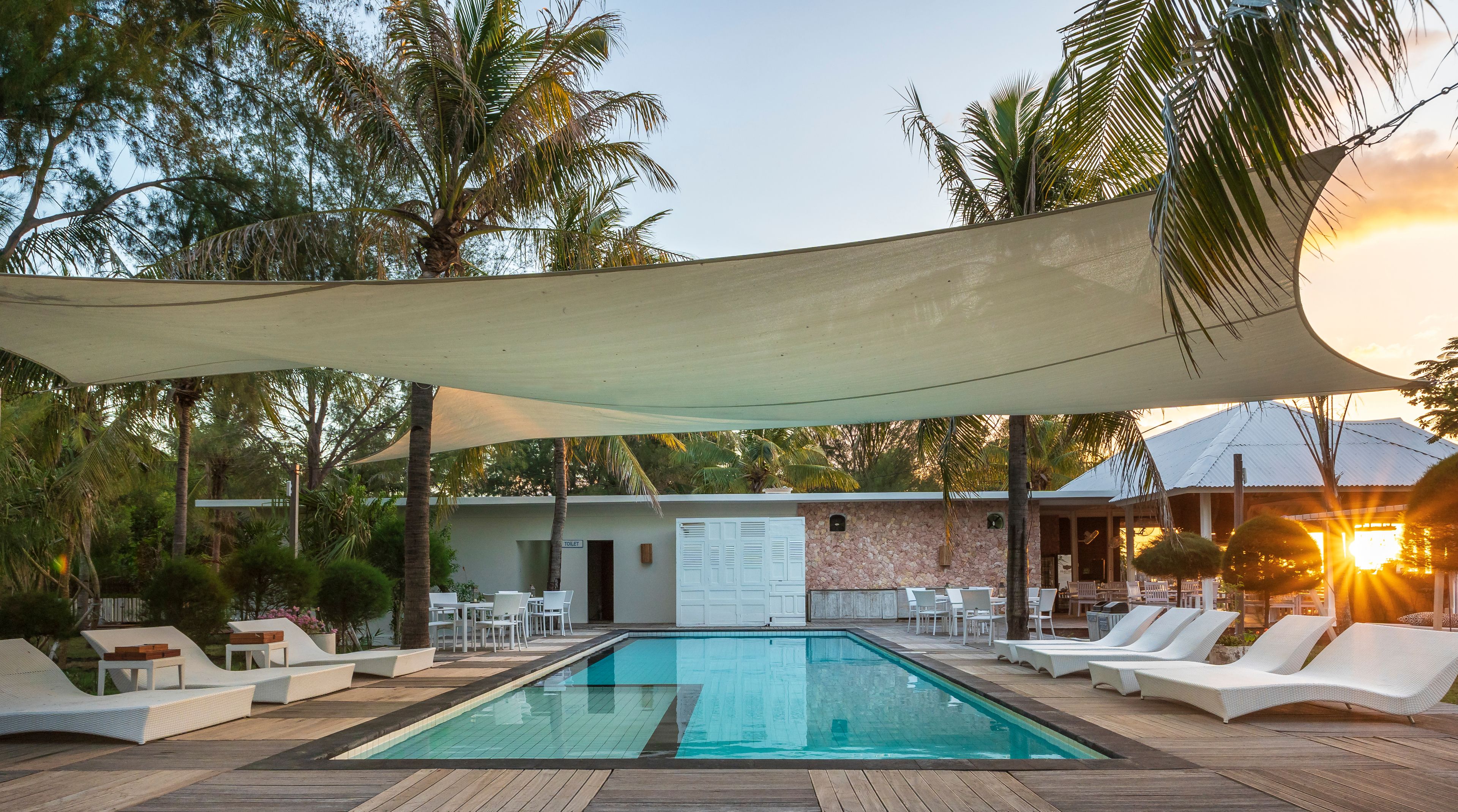Área da piscina com espreguiçadeiras, um espaço coberto para sentar e um edifício com o símbolo de sanitário.