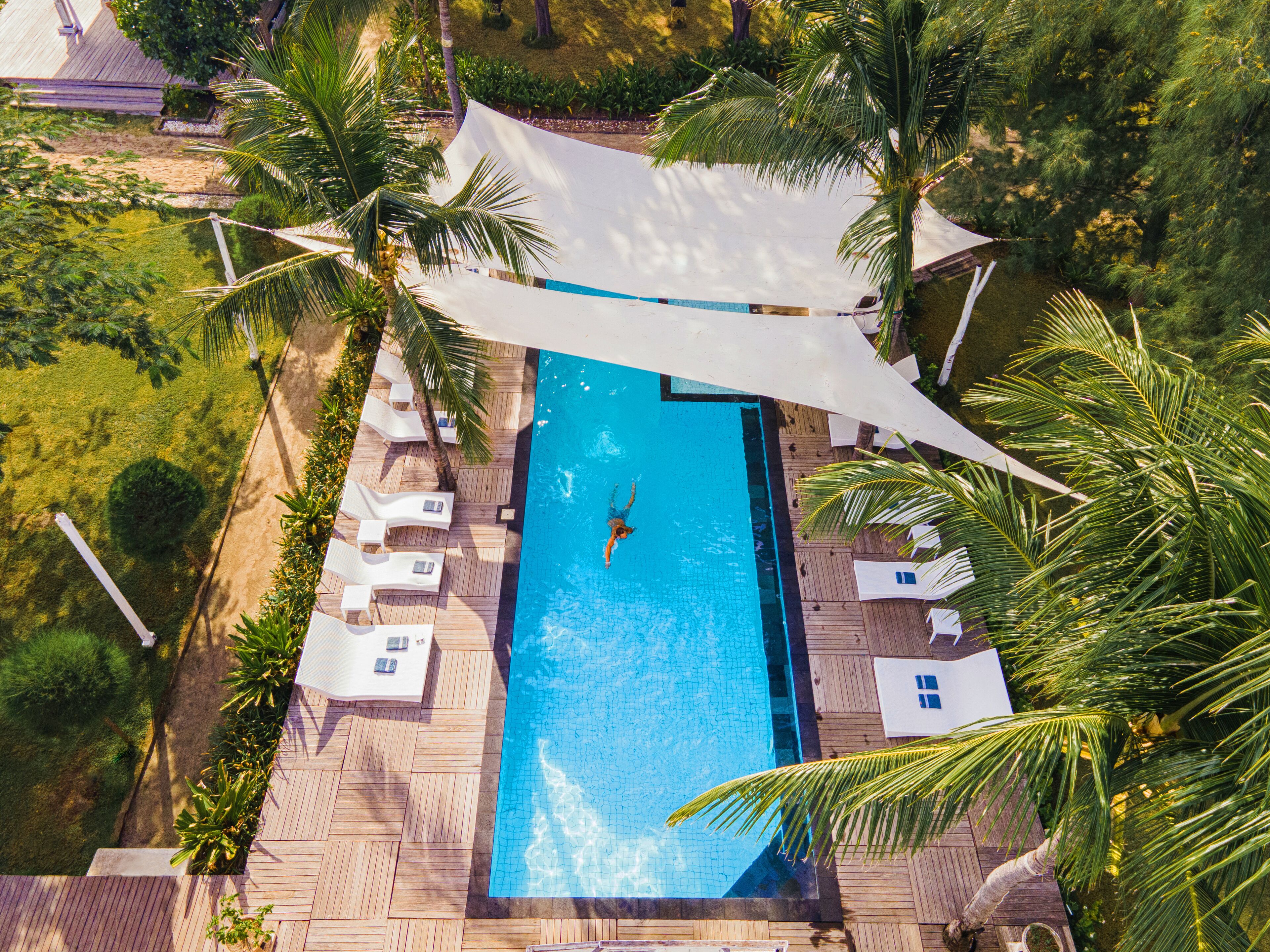 An aerial view of a rectangular swimming pool surrounded by wooden deck, white lounge chairs, and palm trees.