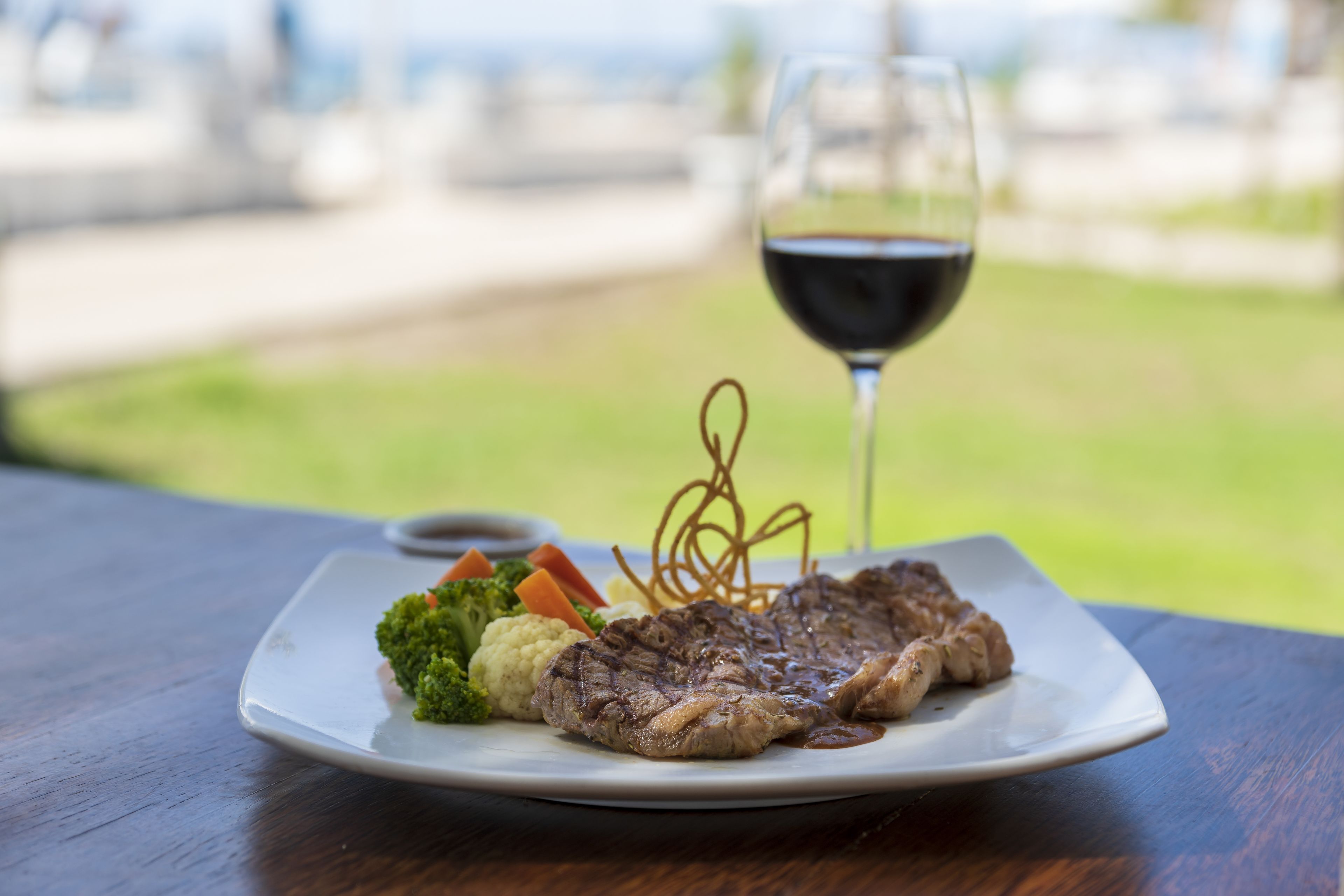 Een bord met steak, groenten en een glas rode wijn op een tafel met uitzicht op een grasveld.