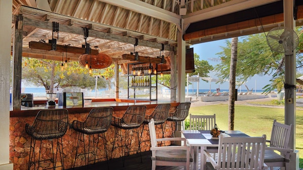 Un espacio de estar al aire libre techado con sillas de mimbre y una mesa, con vistas a una playa con sombrillas y sillas de descanso.