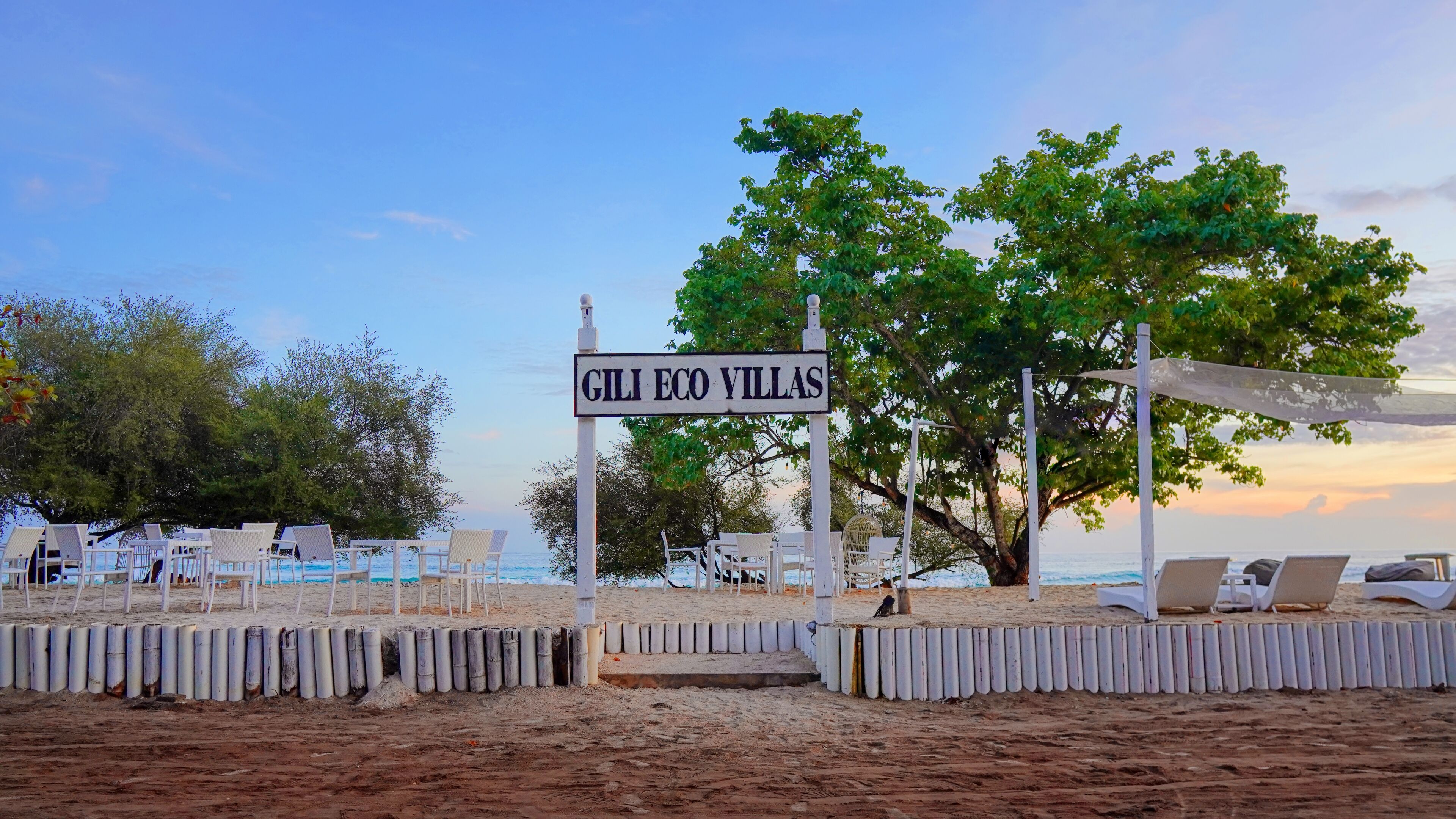 Zona de playa con un cartel que dice "GILI ECO VILLAS", sillas de descanso blancas y un árbol.
