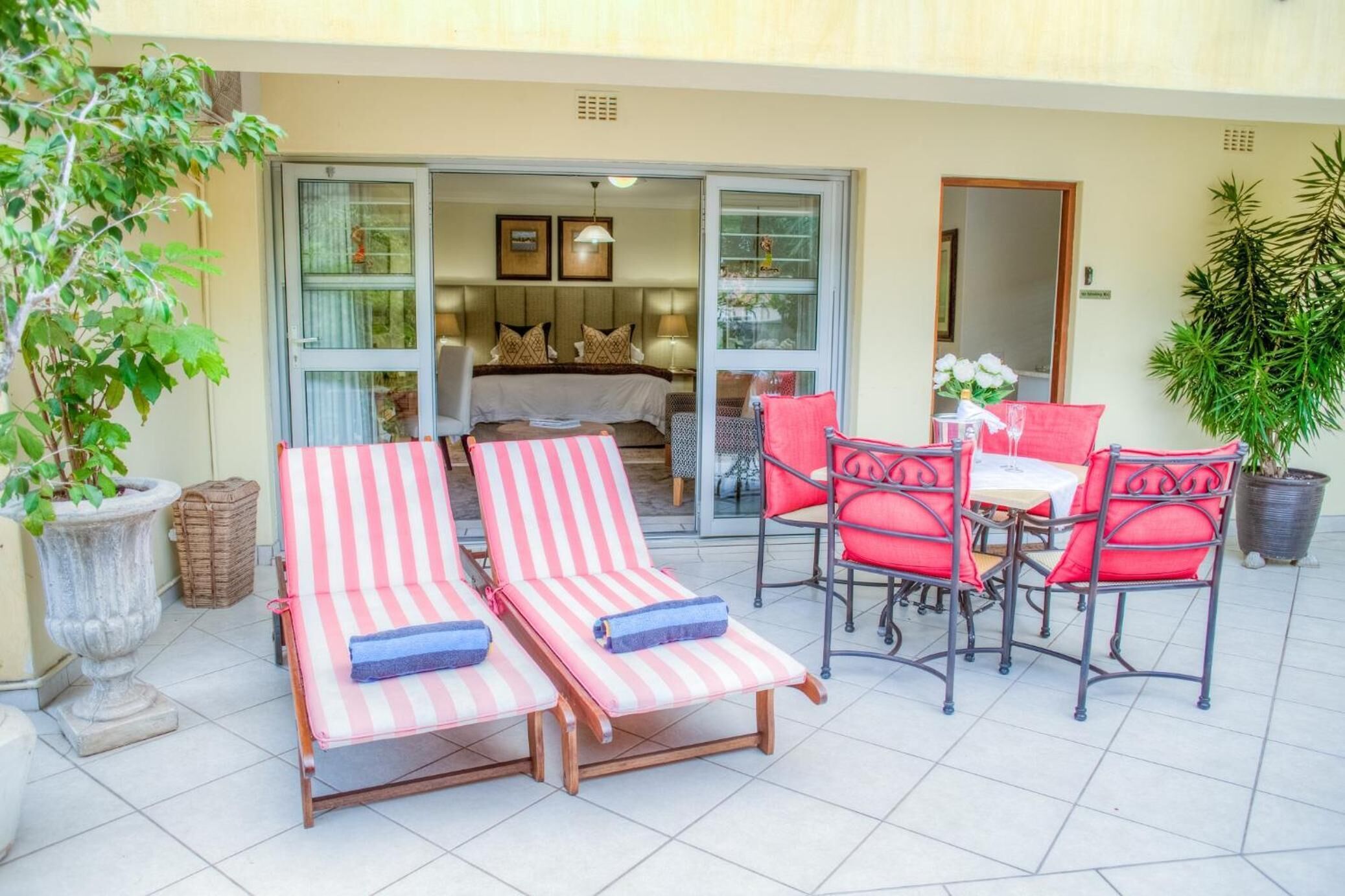 A sunlit patio with striped lounge chairs, a table with red chairs, and a view into a bedroom with a bed and curtains.