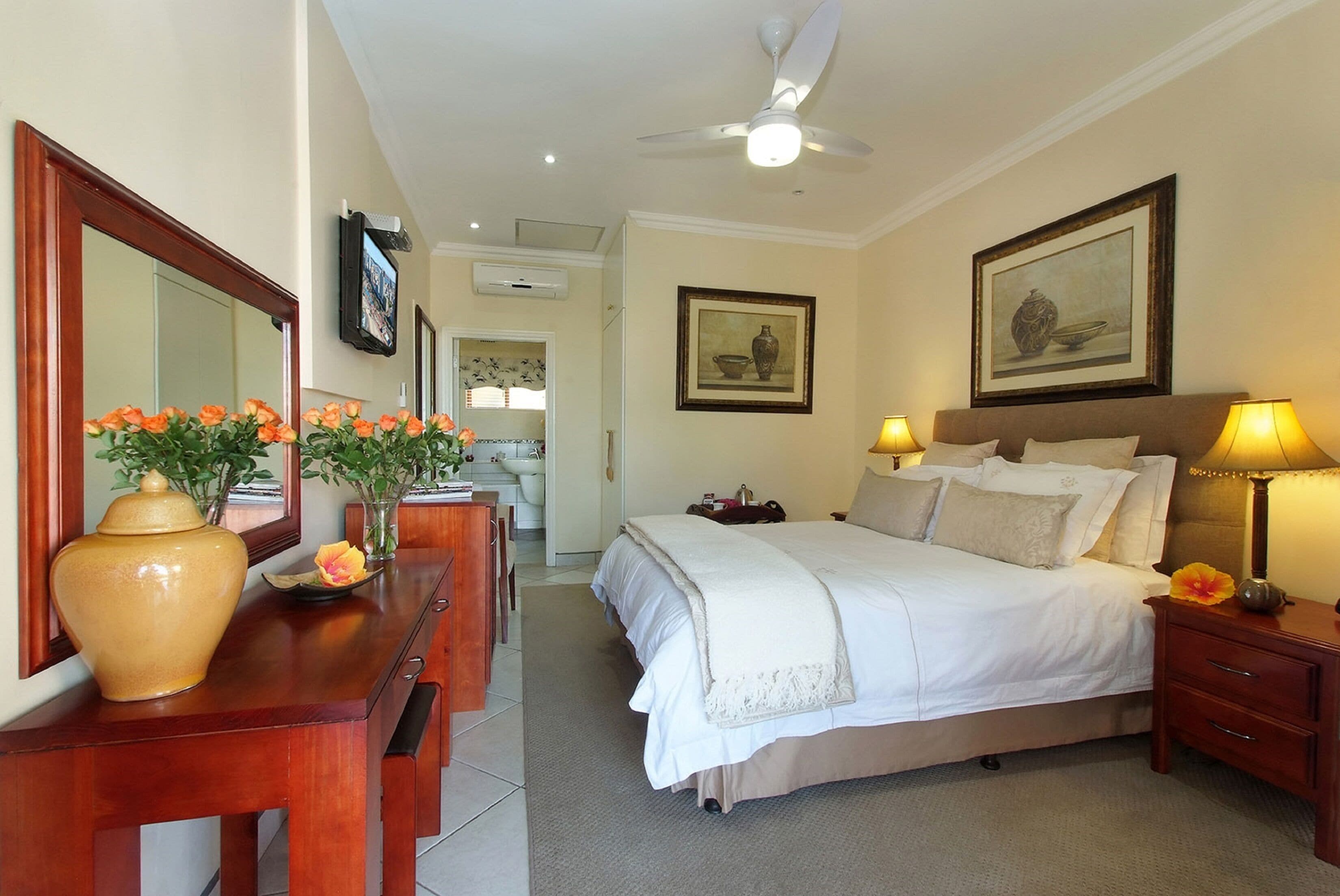 A hotel room with a bed, bedside lamps, a large mirror, a vase with flowers, and a painting on the wall.