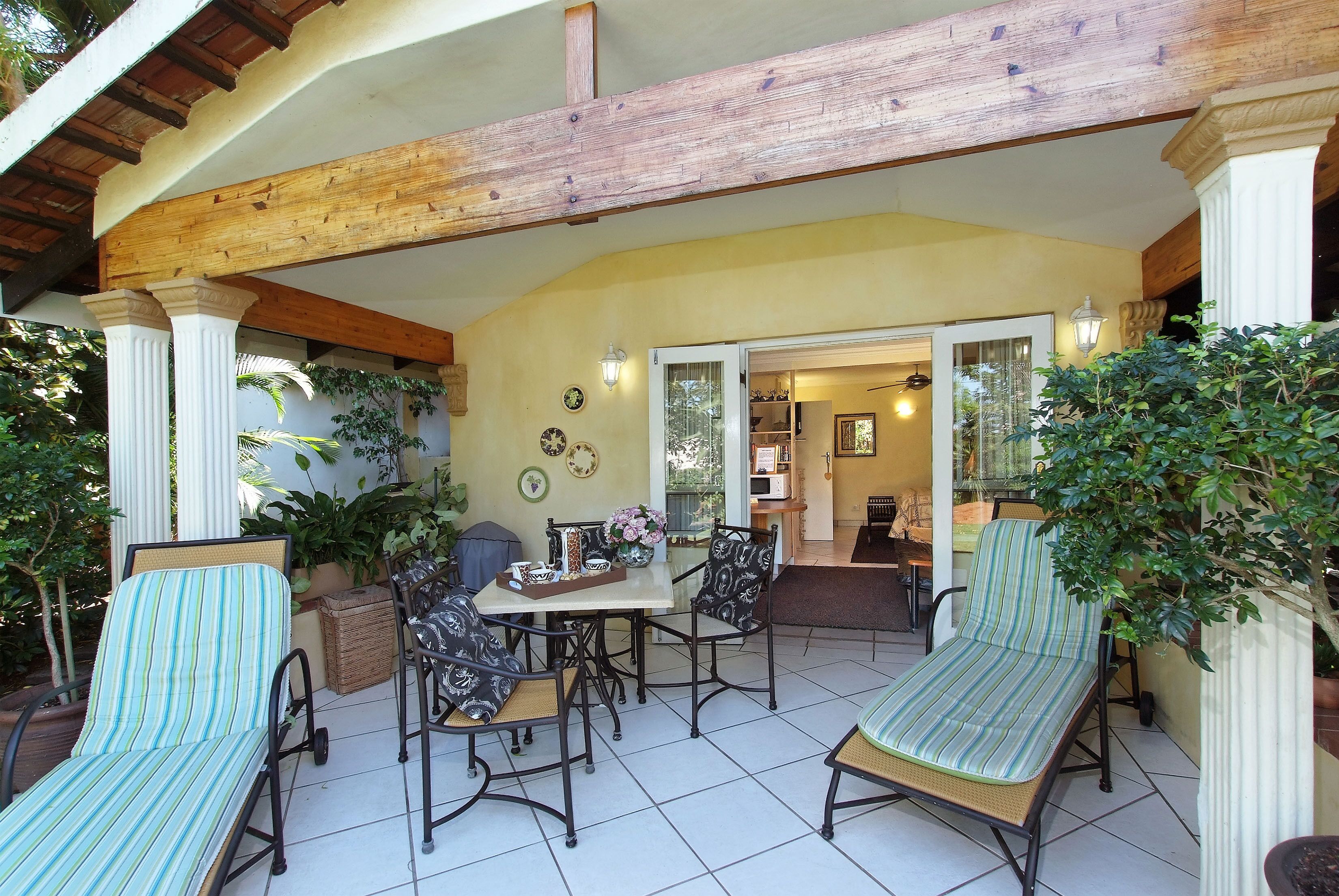 Un área de patio techada con una mesa y sillas, rodeada de plantas en macetas y una estructura de techo de madera.