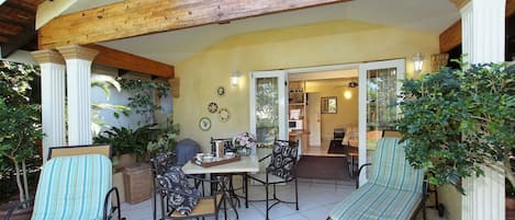 Un área de patio techada con una mesa y sillas, rodeada de plantas en macetas y una estructura de techo de madera.
