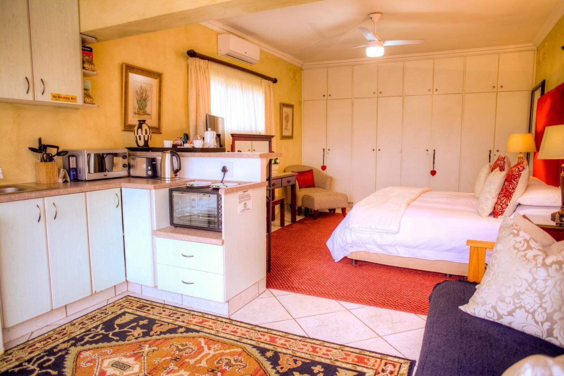 Une chambre d’hôtel compacte comprenant un coin cuisine, un lit avec une literie rouge et blanche, un petit canapé et un tapis rouge à motifs.