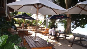 Outdoor wooden picnic tables and umbrellas, with a beach and palm trees in the background.