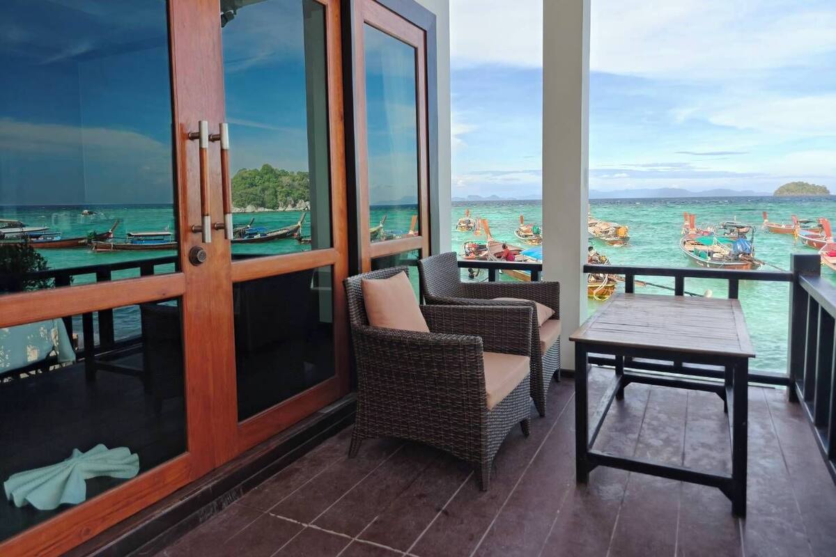 A balcony with wicker furniture and a table, overlooking a sea with boats.