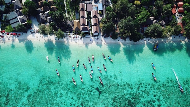 An aerial view of a beach with clear turquoise waters and several small boats. There are buildings with red roofs and umbrellas along the shore. - Cabana Lipe Beach Resort (Koh Lipe)