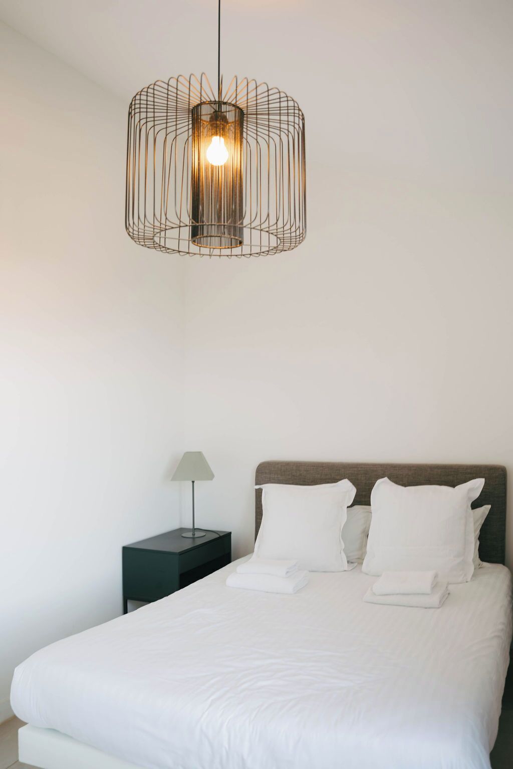 A bedroom with a bed, white pillows, a nightstand with a lamp, and a hanging light fixture.