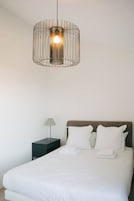 Un dormitorio con cama, almohadas blancas, una mesita de noche con lámpara y una lámpara colgante.