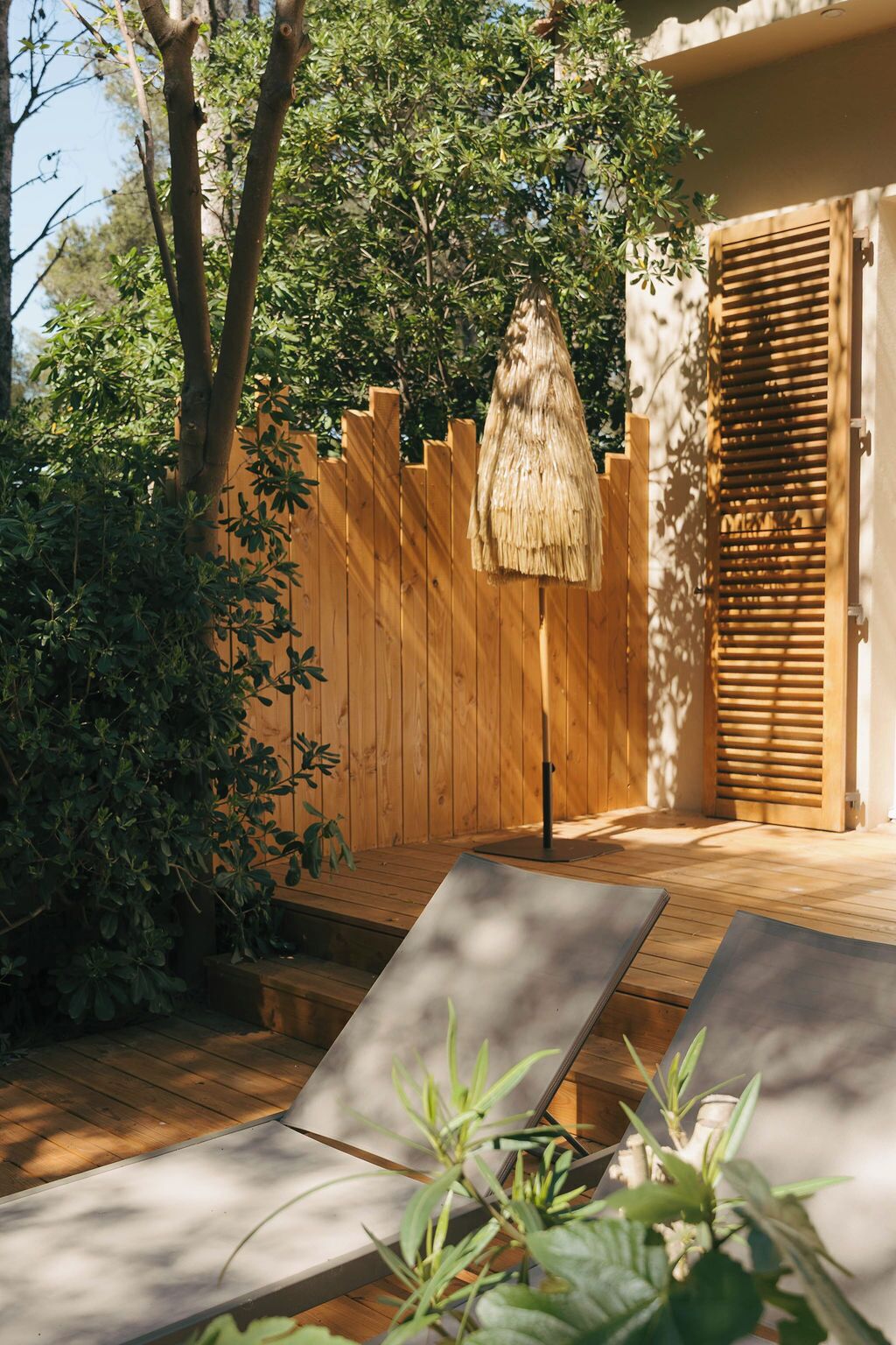 A wooden deck with two lounge chairs, a thatched umbrella, and a wooden fence.