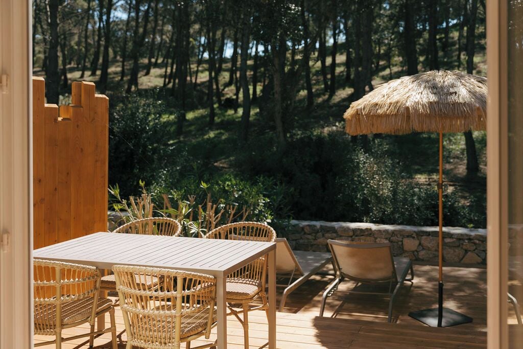 An outdoor patio with a wooden table and wicker chairs, a thatched umbrella, and a stone wall backdrop.