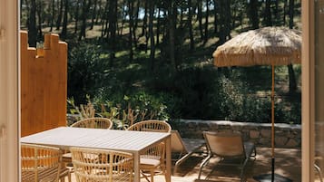 An outdoor patio with a wooden table and wicker chairs, a thatched umbrella, and a stone wall backdrop.