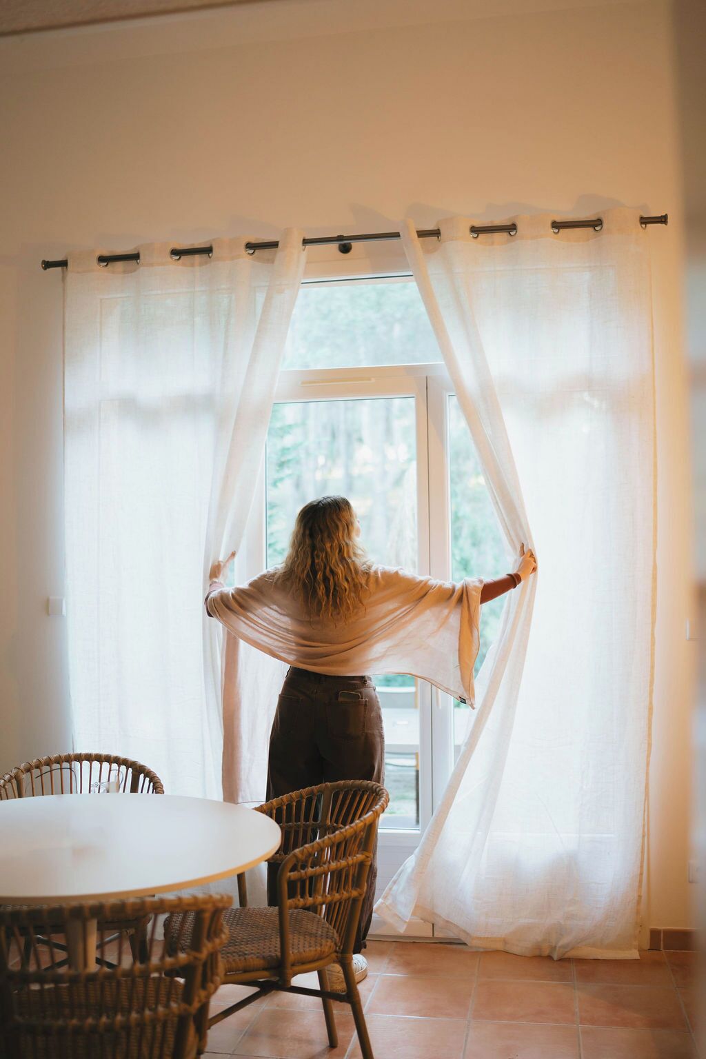 Una persona de pie junto a una ventana con cortinas translúcidas, una silla de mimbre y una mesa en primer plano.