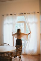 Una persona de pie junto a una ventana con cortinas translúcidas, una silla de mimbre y una mesa en primer plano.