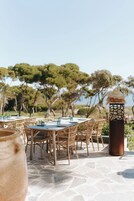 Un espacio de comedor al aire libre con mesas y sillas de mimbre, una maceta metálica decorativa, y vistas a árboles y al cielo.