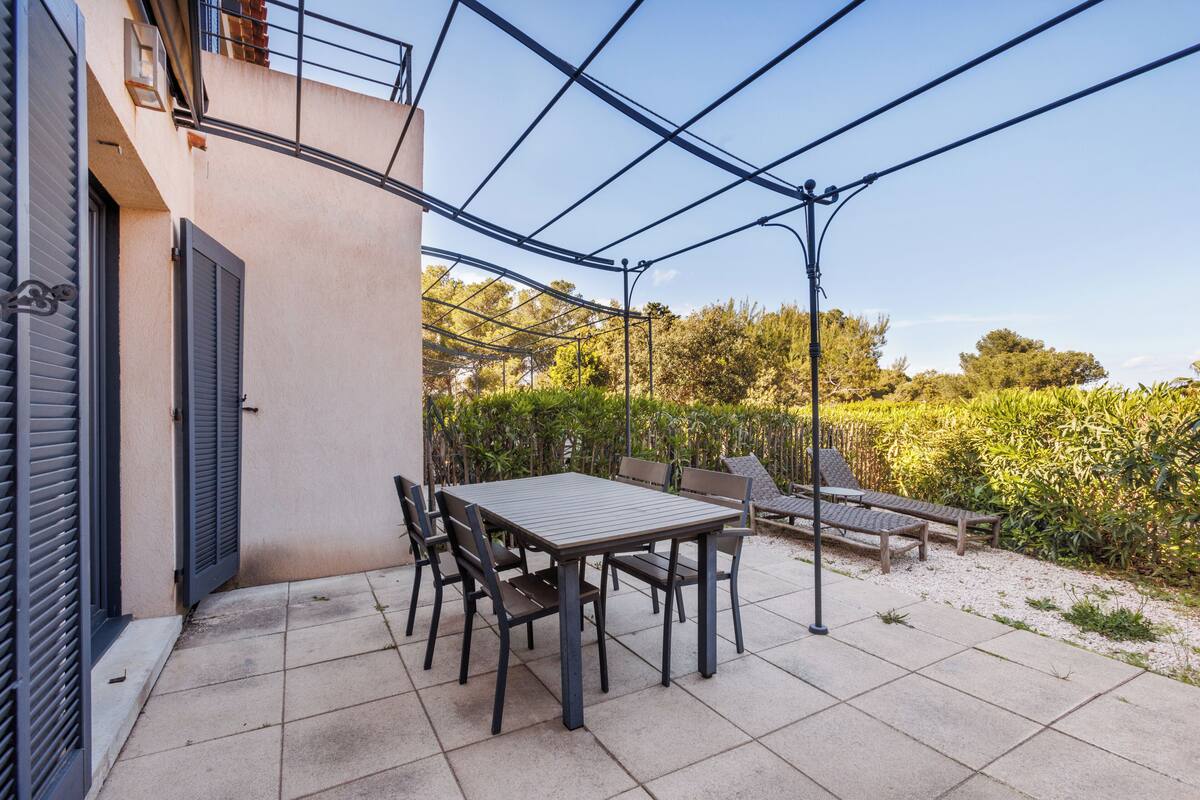 An outdoor patio with a table and chairs, surrounded by a garden and a building with shutters.