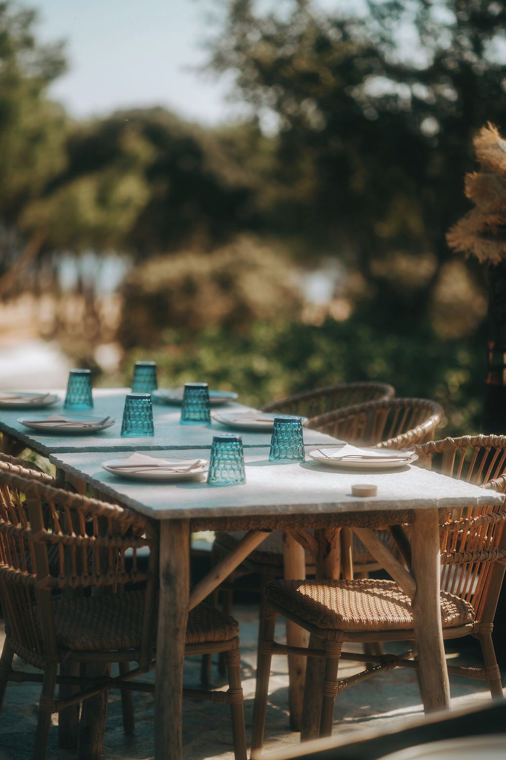 A table set outdoors with wicker chairs, a white tablecloth, and blue glass candle holders.