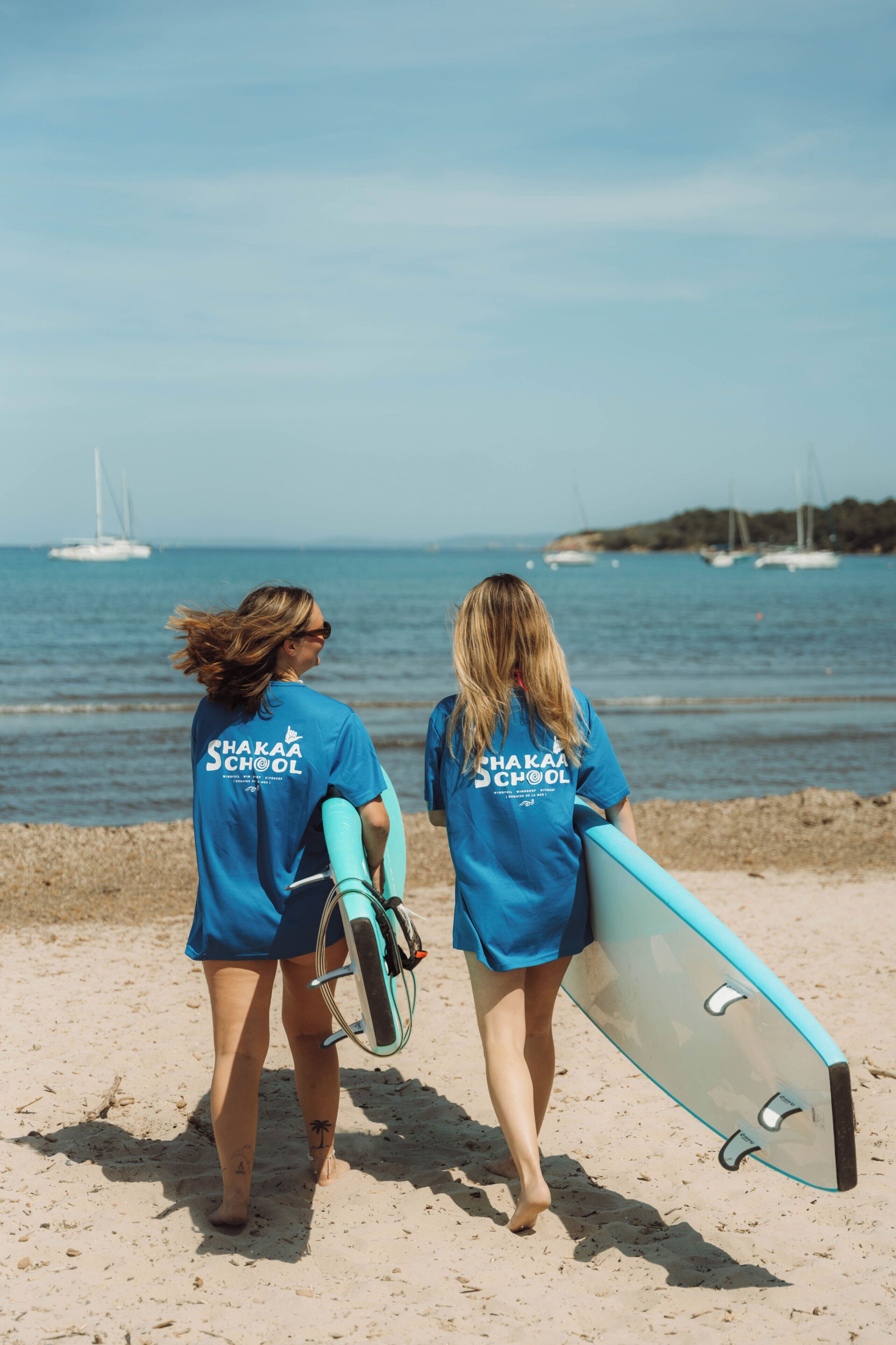 Two individuals walking on a beach with surfboards, wearing blue shirts with 'Shaka School' text, and a clear view of the sea and boats in the background.