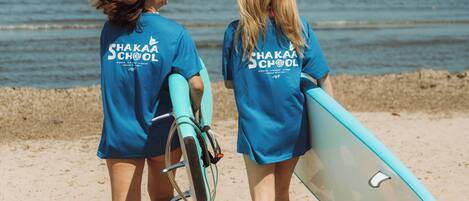 Two individuals walking on a beach with surfboards, wearing blue shirts with 'Shaka School' text, and a clear view of the sea and boats in the background.