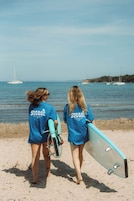 Dos personas caminando sobre una playa con tablas de surf, vistiendo camisetas azules con el texto 'Shaka School' y una vista despejada del mar y barcos al fondo.