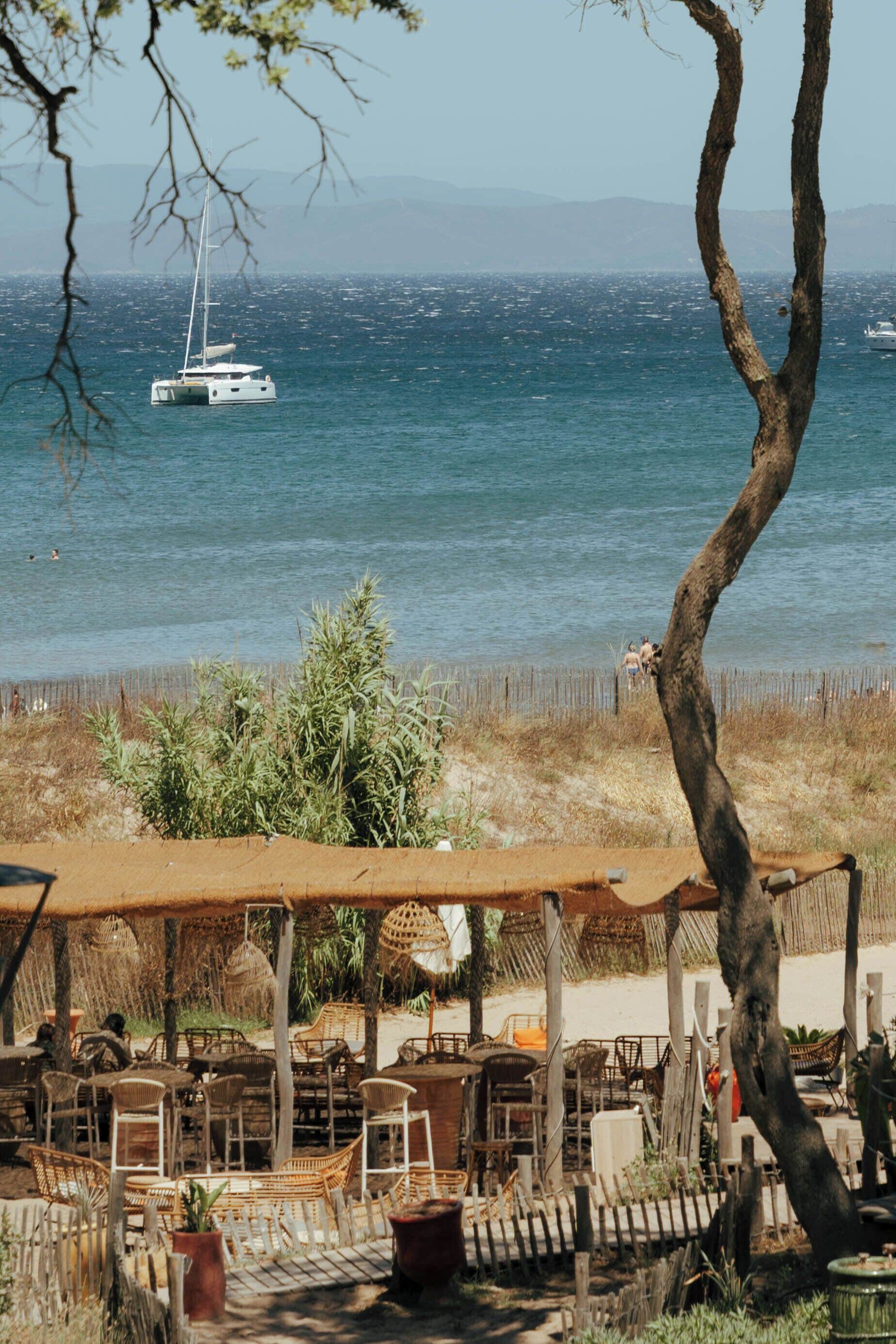 A seaside restaurant with outdoor seating, a view of the sea, and a sailboat in the distance.