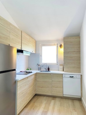 A modern kitchen with wooden cabinets, a stainless steel refrigerator, a built-in dishwasher, and a window providing natural light. - Le Domaine de la Mer (Hyères)