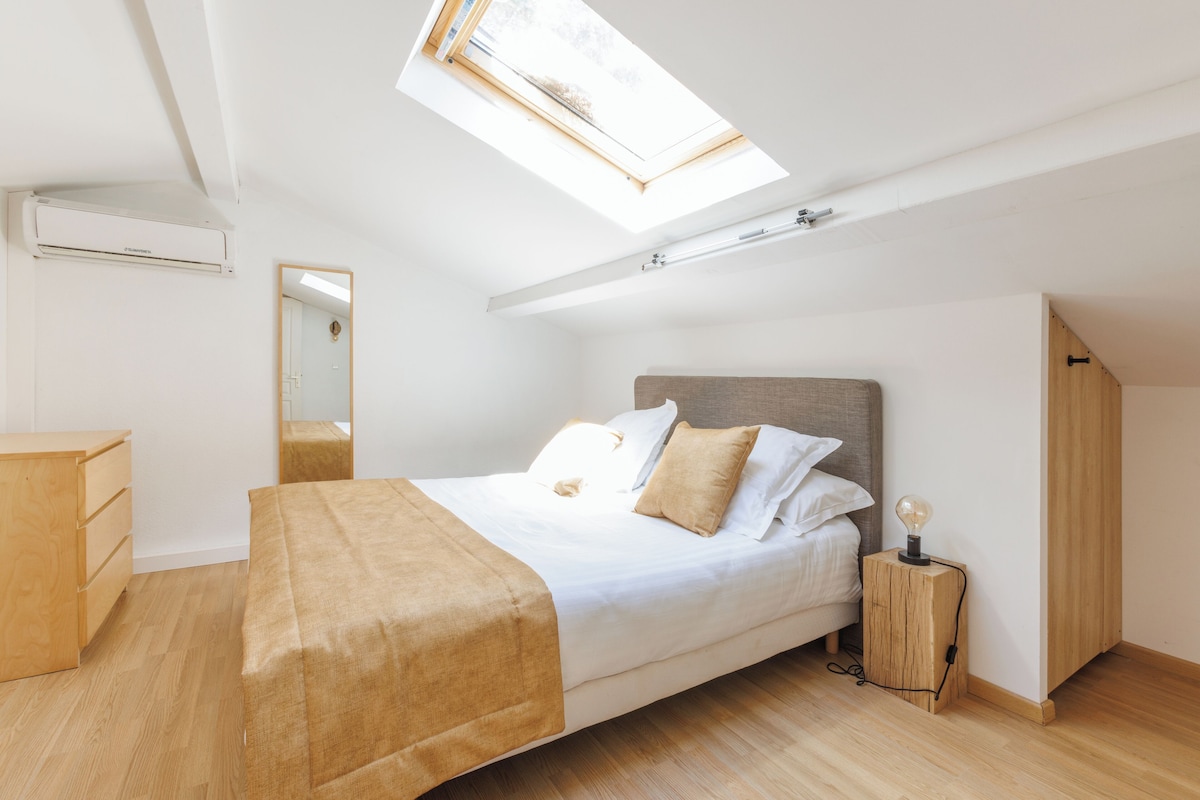 A bedroom with a skylight, a bed with a headboard, a bedside table with a lamp, and a wooden dresser.