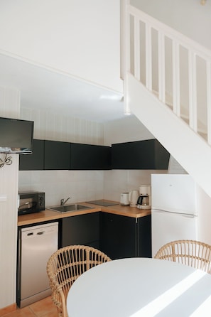 A compact kitchen with a dishwasher, toaster, coffee maker, and a white refrigerator. There is a wicker chair and a small dining table.