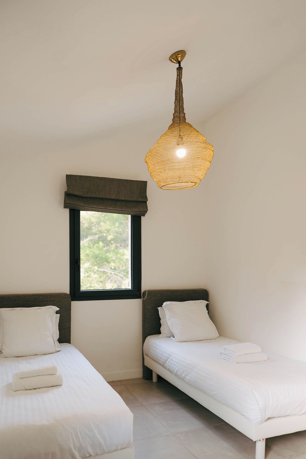 A bedroom with two single beds, a window with a view, and a hanging light fixture.