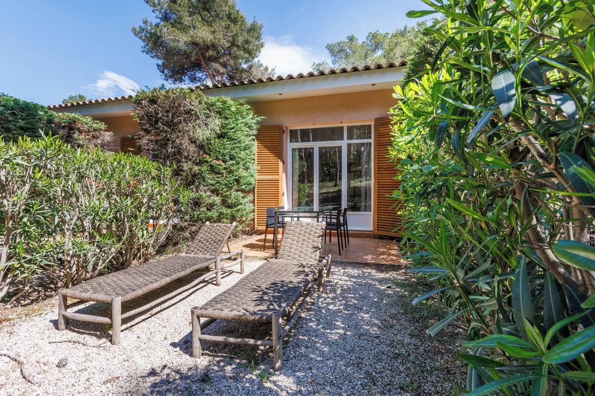 A patio with two wooden lounge chairs and a table, surrounded by gravel and greenery.