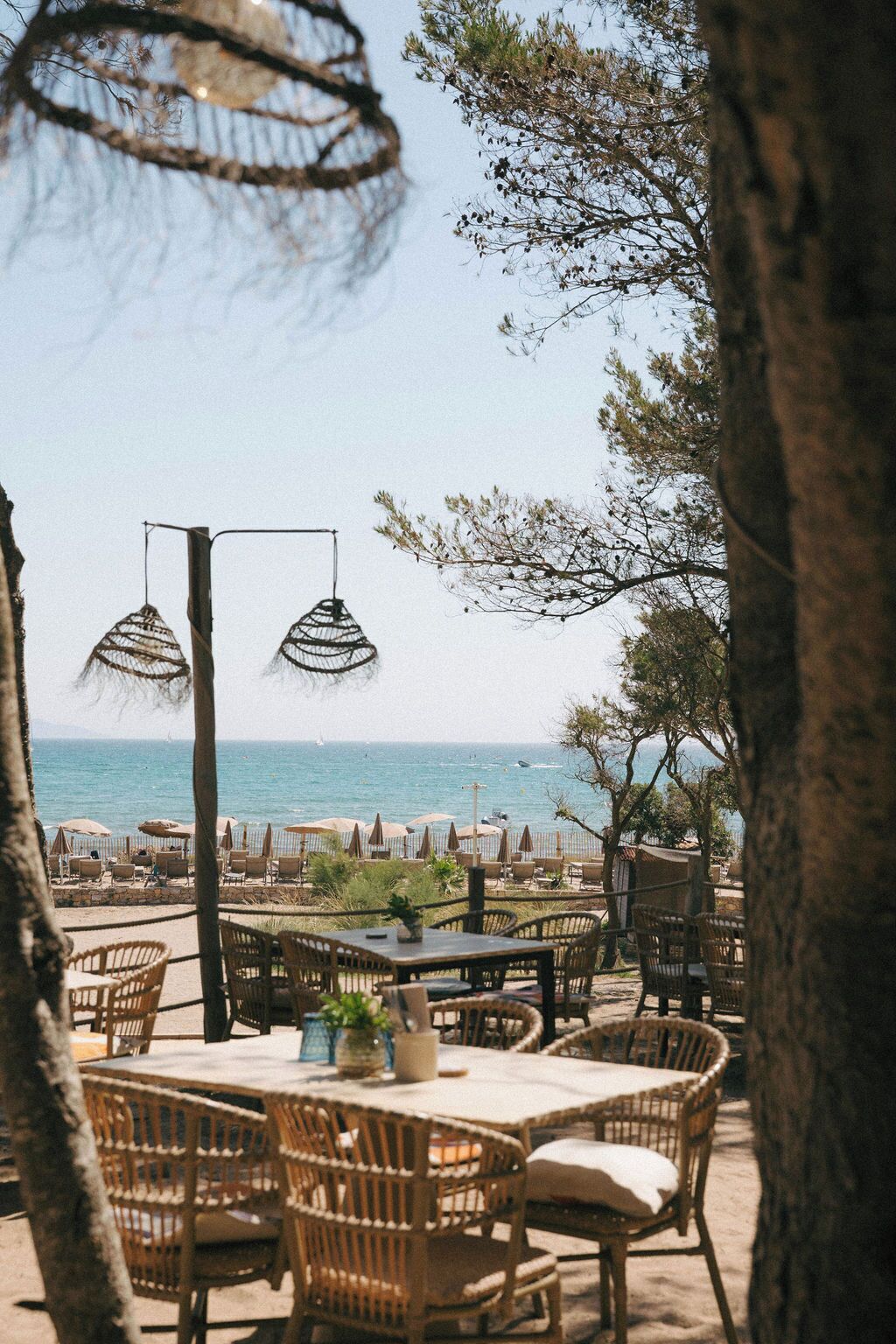 An outdoor seating area with wicker furniture and hanging lights, overlooking a beach with umbrellas and lounge chairs.