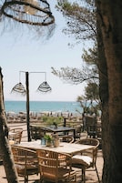 Un espacio al aire libre con mobiliario de mimbre e iluminación colgante, con vistas a una playa con sombrillas y sillas reclinables.