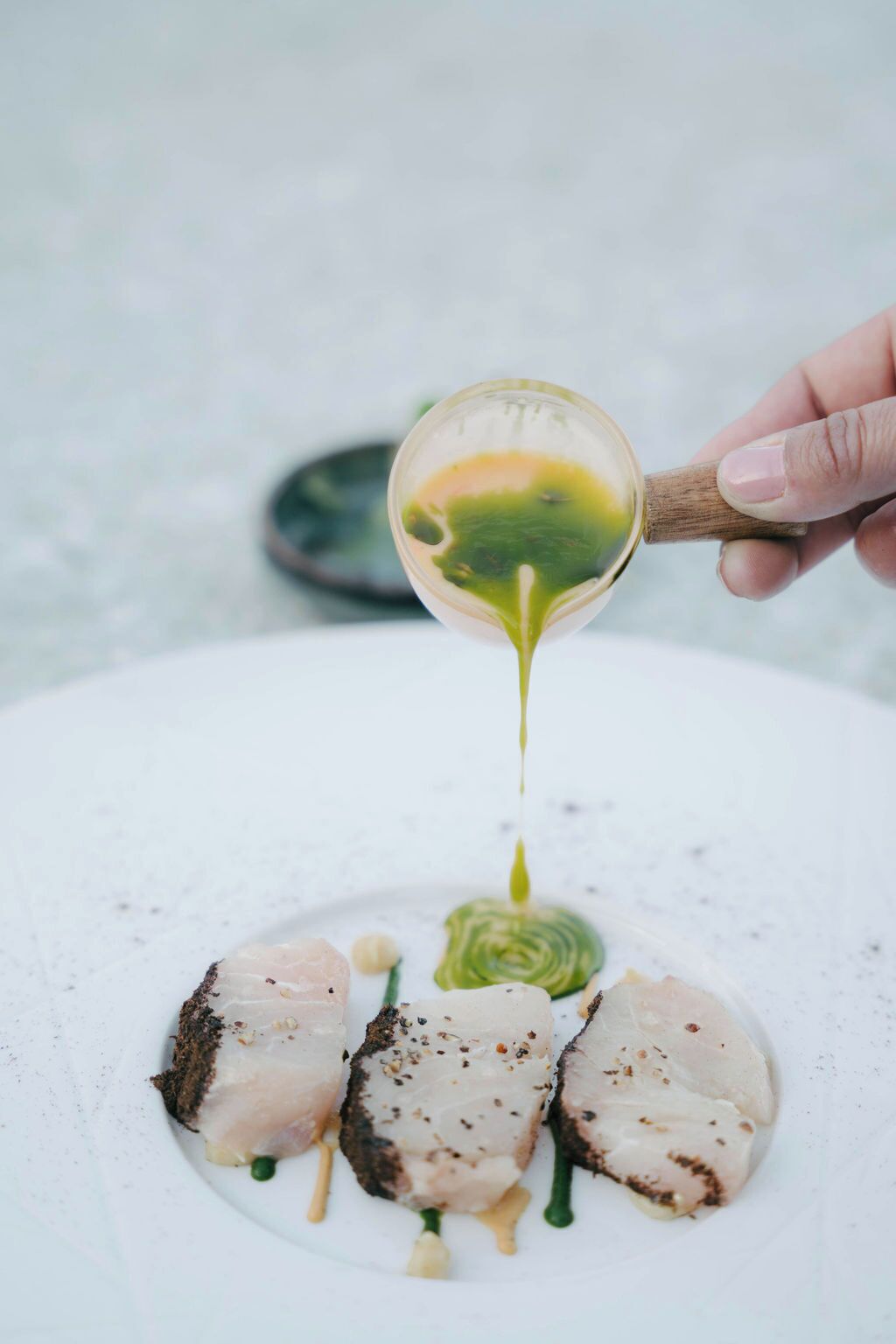 A hand pouring a green sauce over a dish with fish and vegetables.