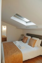 A hotel room with a skylight, a bed with pillows, a bedside table with a lamp, and a mirror on the wall.