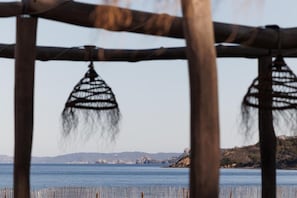 A wooden structure with hanging woven decorations, overlooking a calm body of water with distant landforms. - Le Domaine de la Mer (Hyères)