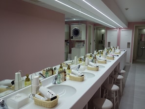 A long, white bathroom with multiple sinks, each with a mirror and various toiletries. There are wicker baskets under the sinks, and the walls are painted pink.