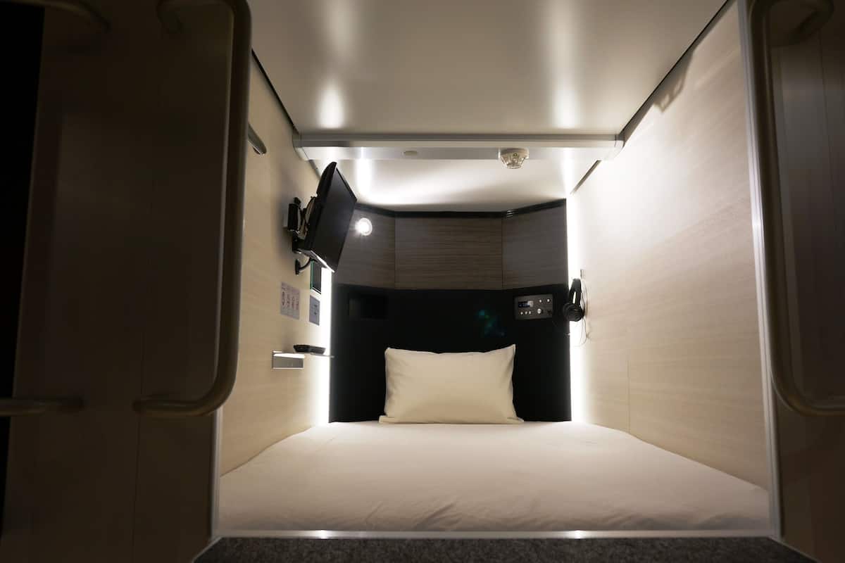 Una habitación estrecha, tenuemente iluminada, con una cama individual, una mesita pequeña y un panel de control en la pared.